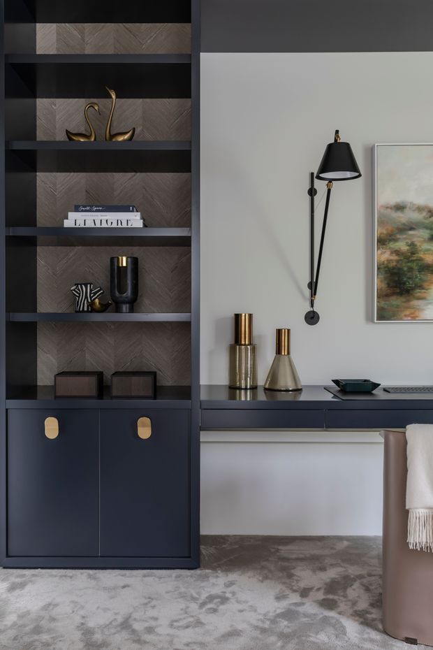 A refined, moody interior vignette featuring a dark built-in shelving and cabinetry unit with herringbone-patterned timber backing, styled with books and decorative objects. A matching dark floating desk or console runs along the pale grey wall beneath a sculptural black wall lamp, metallic vessels, artwork, and part of a taupe upholstered chair. The floor is covered with a large muted grey patterned rug.