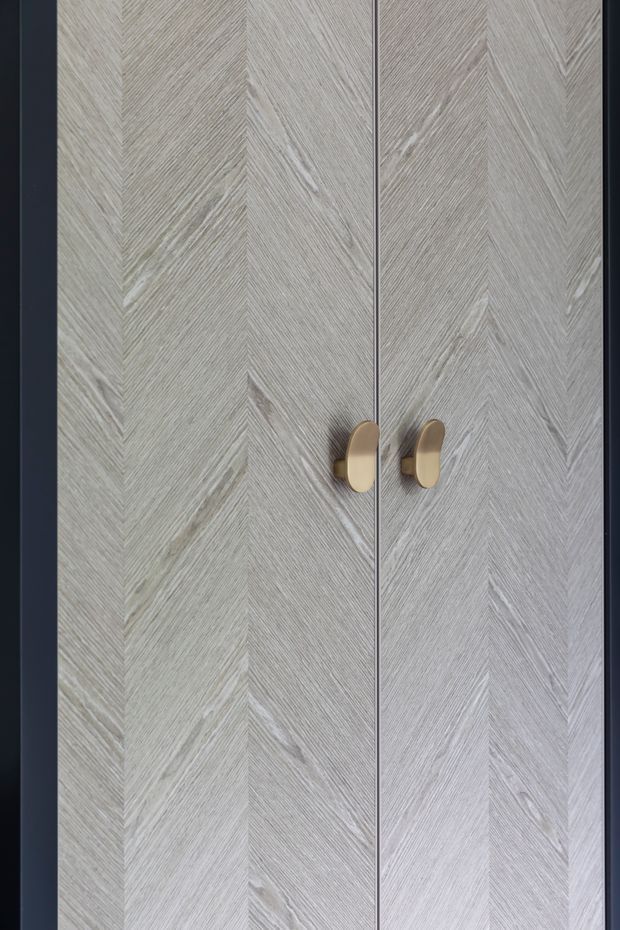 Close-up of tall built-in cabinet doors featuring a pale grey-washed timber veneer arranged in a chevron or herringbone pattern. The doors have slim dark framing and paired rounded brass-gold handles, creating a refined, contemporary joinery detail.