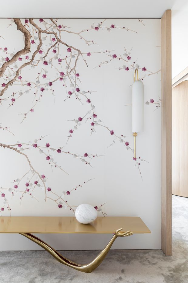 A refined interior vignette featuring a slim light-wood console table with a sculptural polished gold animal-like base, set against a white wall covered in a delicate flowering branch mural. A white cylindrical wall sconce with brass details is mounted on the wall, while a pale stone or ceramic ornament rests on the console. A timber-framed opening reveals a bright adjacent space with light wood cabinetry and pale stone flooring. The room has a soft, elegant, nature-inspired aesthetic.