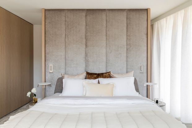 A refined contemporary bedroom with a large upholstered bed set against a full-height vertically channelled grey feature headboard. Warm timber wall panelling and slim timber framing contrast with crisp white bedding, sheer floor-to-ceiling curtains, and minimalist bedside tables and lamps. The room feels calm, bright, and luxurious.