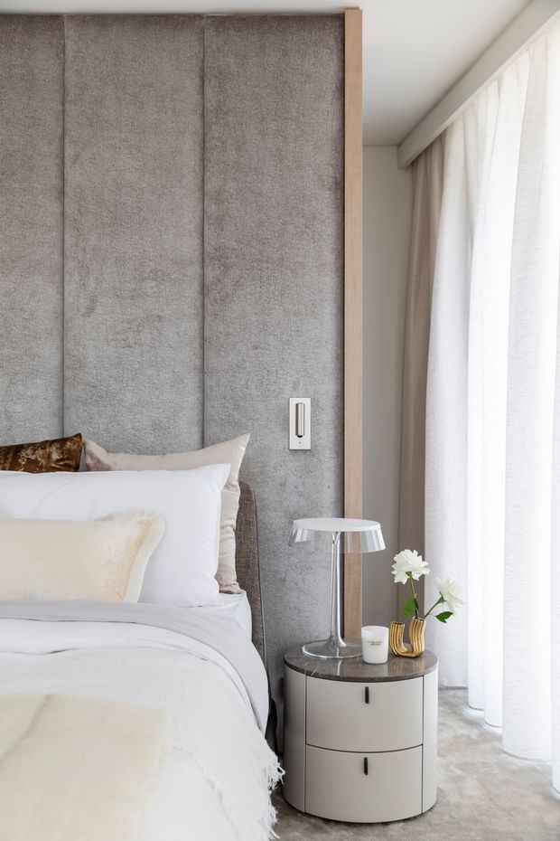 A softly lit contemporary bedroom corner featuring a bed with layered white bedding, a tall upholstered grey feature wall behind the bed, pale timber trim, full-height sheer curtains, and a rounded bedside table with a modern lamp and floral arrangement.