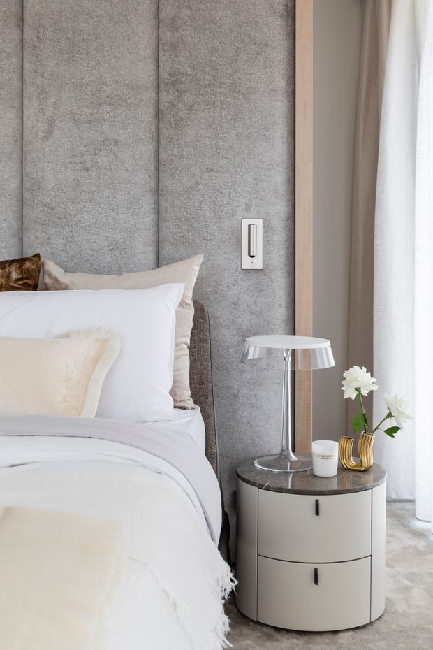 A refined bedroom vignette featuring a bed with layered white and cream bedding against a tall grey upholstered panelled headboard. A rounded two-drawer bedside table with a dark stone top sits beside the bed, styled with a chrome-and-white table lamp, a small cup, and gold-toned vases with white flowers. Pale timber trim and sheer curtains frame the scene, creating a soft, contemporary neutral interior.