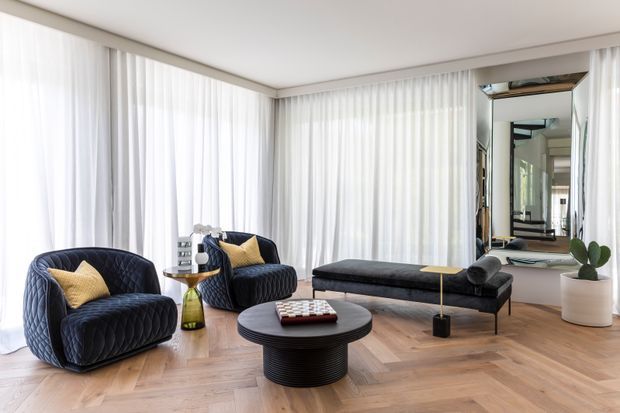 Bright, refined living room with floor-to-ceiling sheer curtains, a large mirrored wall, herringbone timber flooring, two deep navy upholstered armchairs, a matching low chaise-style sofa, and a round black ribbed coffee table. A sculptural floor lamp and potted plant add understated accents.