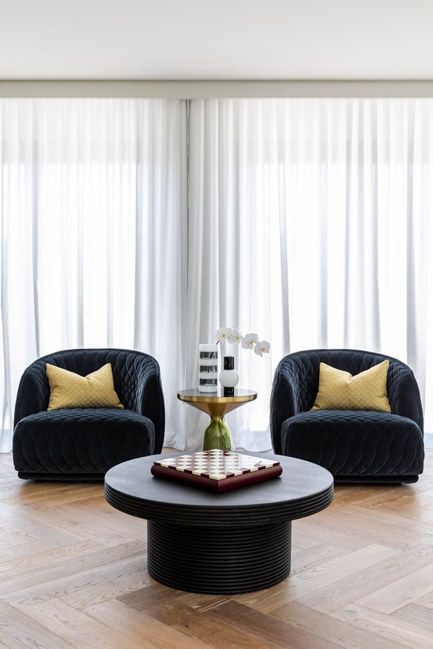 A refined living room with two deep navy quilted lounge chairs, mustard accent cushions, and a round black ribbed coffee table topped with a decorative chessboard. Sheer white curtains span the full-height glazing, while a small brass-and-glass side table and floral arrangement sit between the chairs. The room has warm herringbone timber flooring and a high-contrast, glamorous contemporary aesthetic.