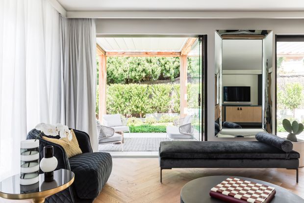 Bright contemporary living room with herringbone timber flooring, a black tufted lounge chair, matching chaise-style sofa, round coffee table and decorative objects. Full-height glazing and sliding doors open to a covered outdoor terrace and landscaped garden, while a mirrored wall panel reflects a television and cabinetry.