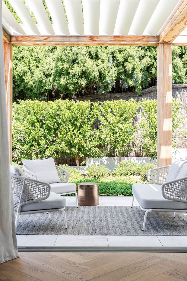 Bright outdoor lounge or covered terrace viewed from inside, with a white slatted pergola roof supported by natural timber beams, pale upholstered lounge chairs, a grey area rug, and a small metallic side table. Lush clipped hedges and mature greenery create a private garden backdrop, while timber herringbone flooring transitions to large pale terrace tiles.