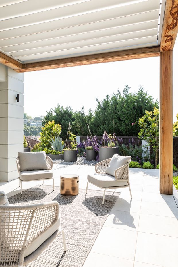 A bright covered outdoor terrace with large white floor tiles, timber posts and beams, and a white louvred roof. Woven outdoor lounge chairs with light cushions surround a small metallic cylindrical table on a textured neutral rug. Potted agave and flowering plants line the edge, with dense greenery and a residential hillside visible beyond.
