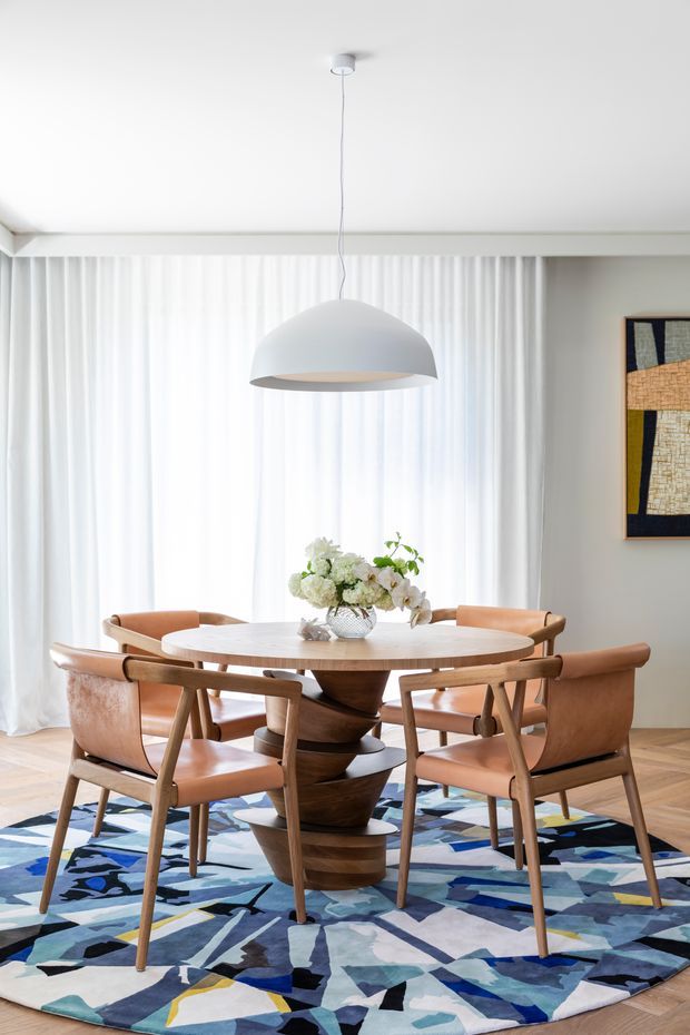 Bright, refined dining room featuring a round light-wood dining table with a sculptural dark-wood pedestal base, surrounded by mid-century-inspired tan leather and timber chairs. A large white dome pendant hangs centrally above the table, with full-height sheer white curtains, pale walls, timber flooring, and a bold blue, white, grey, and black abstract rug. A framed textile artwork adds visual interest on the wall.