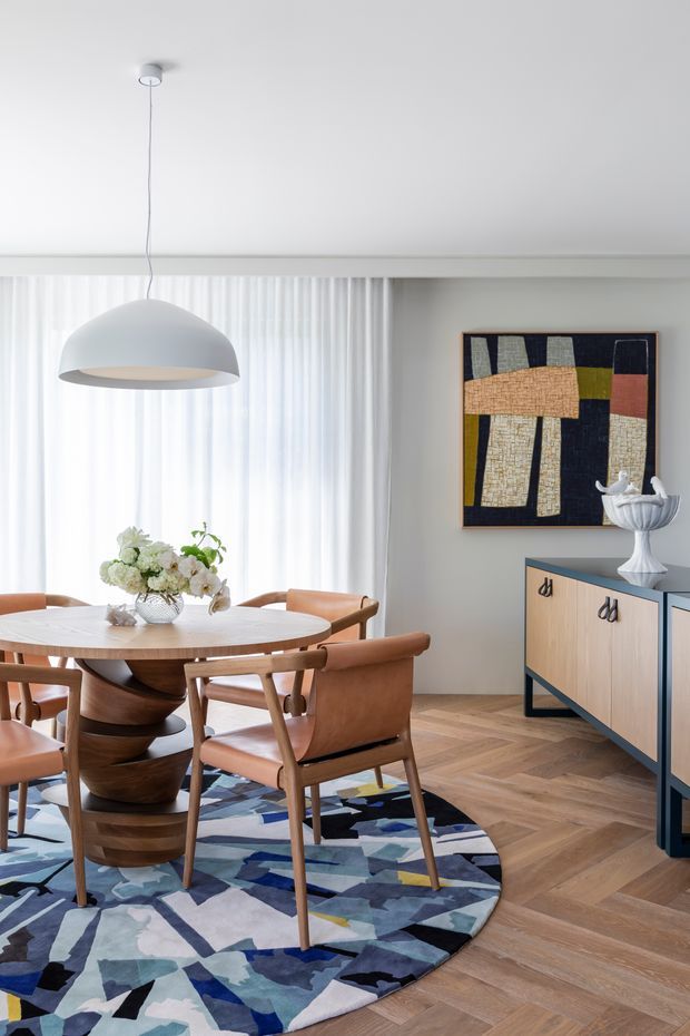Bright, refined dining room with a round timber dining table and sculptural pedestal base surrounded by upholstered timber dining chairs. A large white dome pendant hangs above the table, with sheer floor-to-ceiling curtains, a colourful abstract artwork, a blue-framed timber sideboard, decorative ceramics, and a bold geometric area rug over herringbone timber flooring.