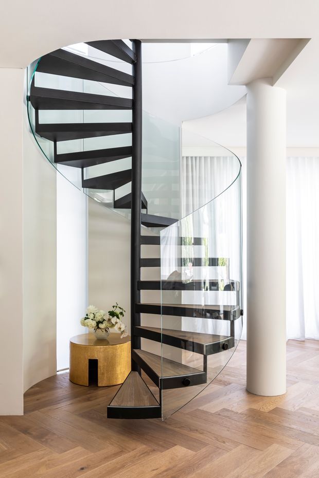 Bright contemporary interior featuring a sculptural spiral staircase with dark timber or metal treads, a central black structural spine, and curved transparent glass balustrades. The staircase is set against white walls and cylindrical columns, with warm herringbone timber flooring, sheer curtains, and a small gold-toned side table with flowers.