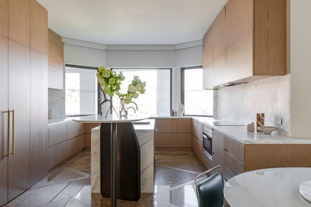 A bright, refined contemporary kitchen with extensive warm timber cabinetry, pale stone benchtops and splashback, and a central island with dark contrasting sculptural sides. The kitchen has a broad bay of windows, polished stone flooring, integrated appliances, and a partial dining area in the foreground.
