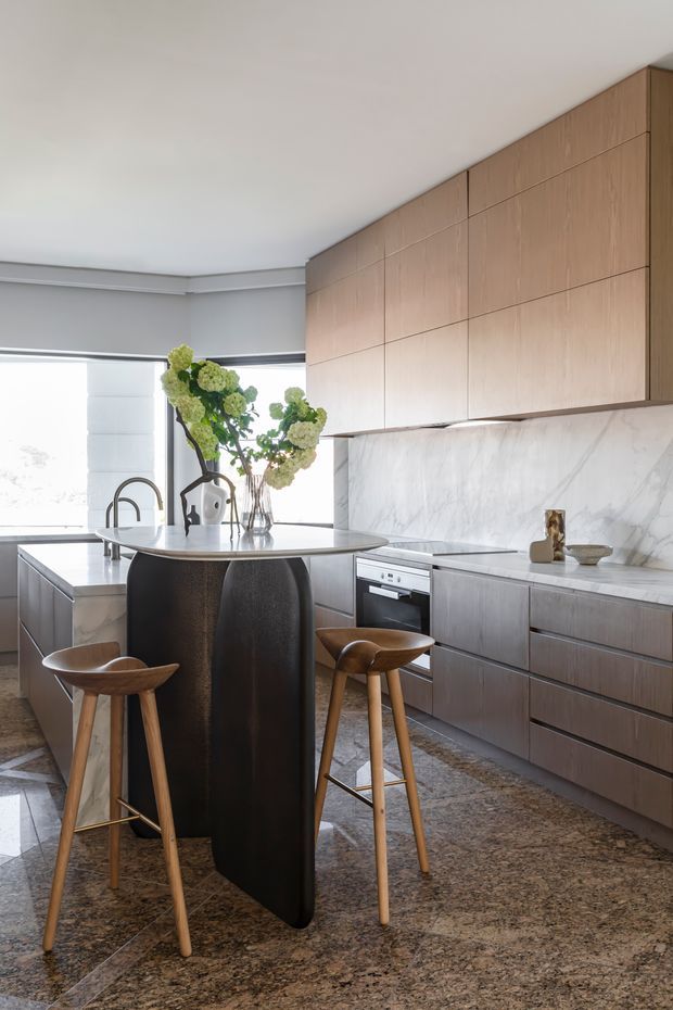 A refined contemporary kitchen featuring a sculptural dark island with timber bar stools, extensive warm timber cabinetry, a pale veined stone splashback and benchtops, integrated appliances, and large windows with roller blinds. The space has a polished, high-end appearance with natural floral styling and a patterned stone floor.