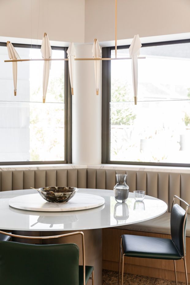 Bright, refined dining nook with a curved upholstered banquette beneath large corner windows. A glossy white oval table, sculptural glassware, dark green and black dining chairs with slim brass-toned frames, and a linear pendant holding folded white shades create a contemporary, softly luxurious interior.