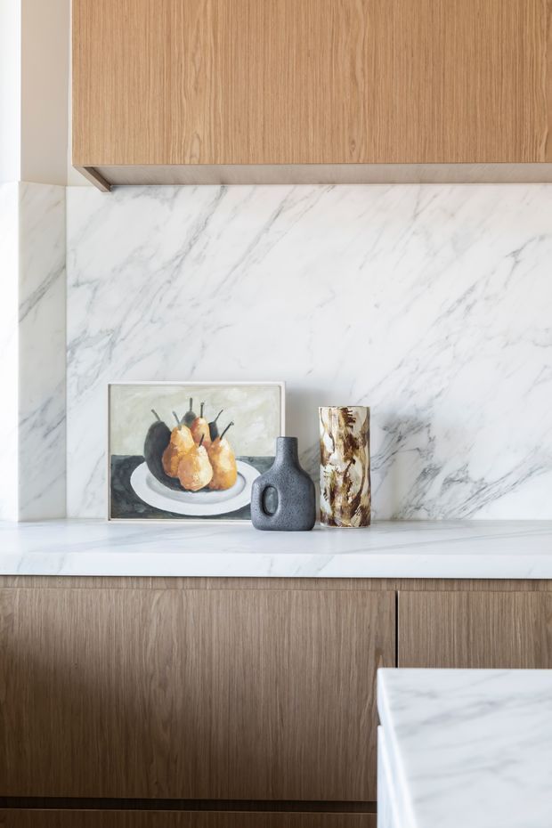 Close-up of a refined kitchen or kitchenette joinery wall featuring warm vertical-grain timber cabinetry, a white marble backsplash and counter with grey veining, and a small still-life arrangement of vases and artwork. The composition is bright, minimal, and contemporary with natural materials and understated styling.