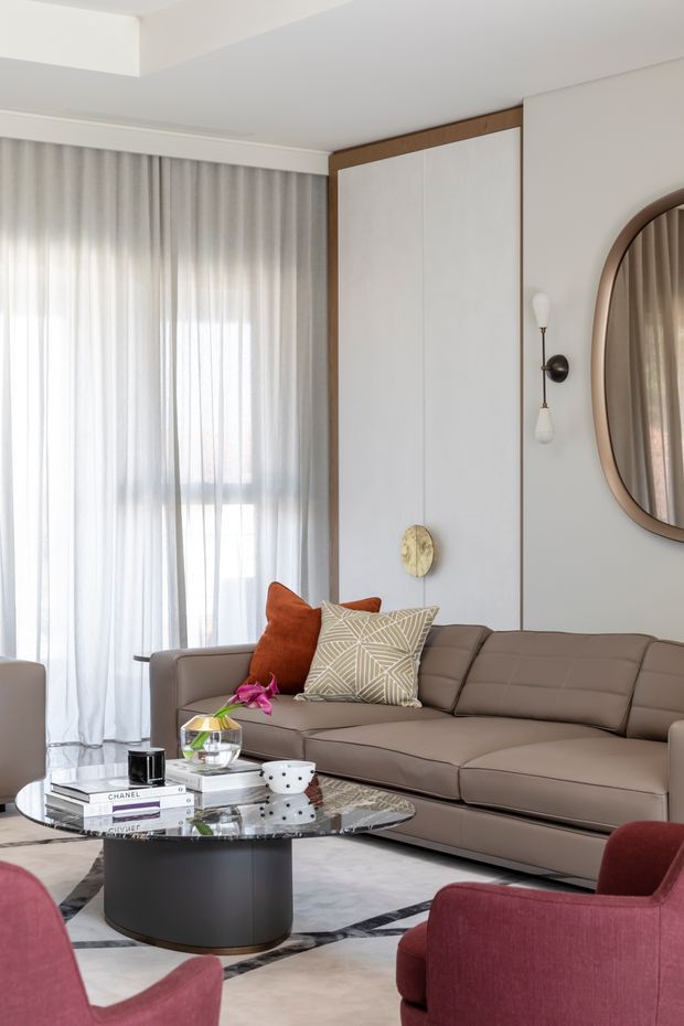 A refined contemporary living room with a large taupe sectional sofa, burgundy upholstered armchairs, a round black coffee table with a glass top, patterned and rust-coloured cushions, sheer floor-to-ceiling curtains, and a large rounded wall mirror. White built-in wall panelling is framed with warm timber trim, complemented by sculptural wall lighting and a pale rug with dark geometric edging.