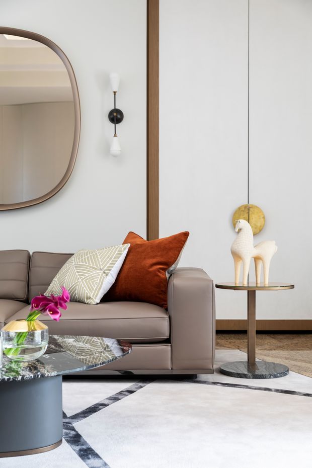 A refined living room vignette featuring a taupe upholstered sofa, patterned and rust-orange cushions, a large rounded rectangular mirror, a sculptural wall sconce, and a slim side table with decorative objects. A glossy dark coffee table, floral arrangement, and geometric area rug complete the contemporary setting. The room has white walls with dark timber trim and a calm, sophisticated palette.