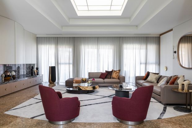 A refined contemporary living room with a large sectional sofa, burgundy lounge chairs, layered coffee tables, full-height sheer curtains, a marble or stone media wall, and a large geometric area rug. A skylight provides abundant natural light, while curved cabinetry and a rounded wall mirror add sculptural detail.