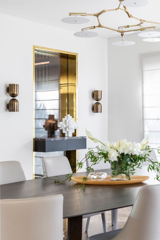 Elegant contemporary dining room with a dark wood dining table, upholstered light chairs, sculptural floral centrepiece, brass-framed mirrored wall feature, bronze wall sconces and a statement branching brass ceiling light. The room is predominantly white with warm metallic accents and refined decorative styling.