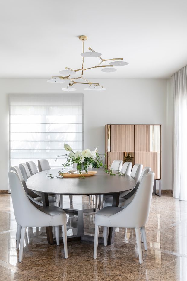 Bright, refined dining room with a large round dark dining table surrounded by upholstered white dining chairs. A sculptural brass-and-white multi-arm pendant light hangs above, while sheer window coverings provide abundant natural light. The room features glossy brown stone flooring, pale walls, floor-to-ceiling curtains, and a built-in wood-panelled display cabinet with brass framing.