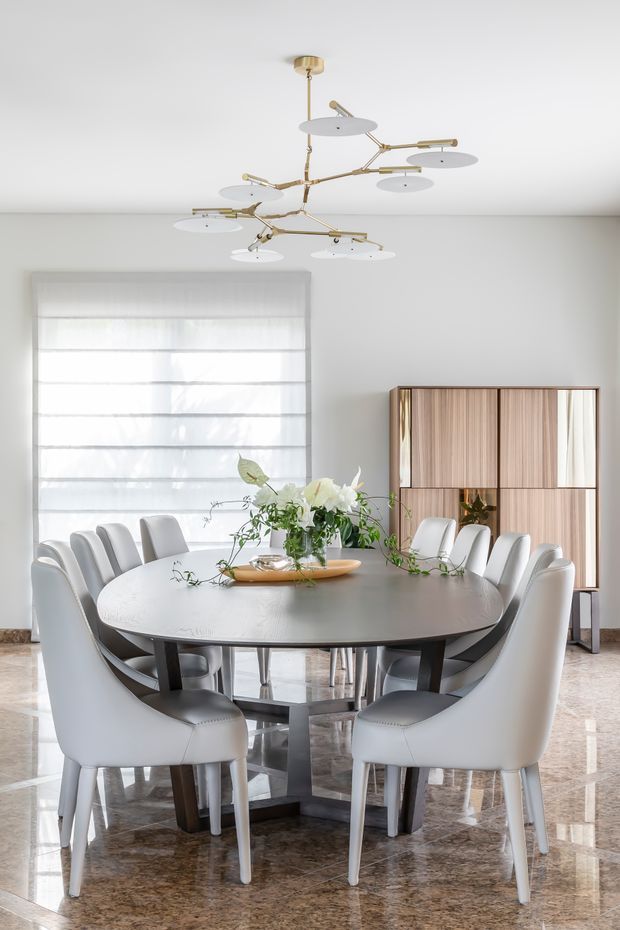 Bright, refined dining room featuring a large oval dark dining table surrounded by upholstered white dining chairs, a sculptural brass and white multi-arm pendant light, sheer Roman blinds, a timber and glass display cabinet, floral table arrangement, and polished stone flooring.