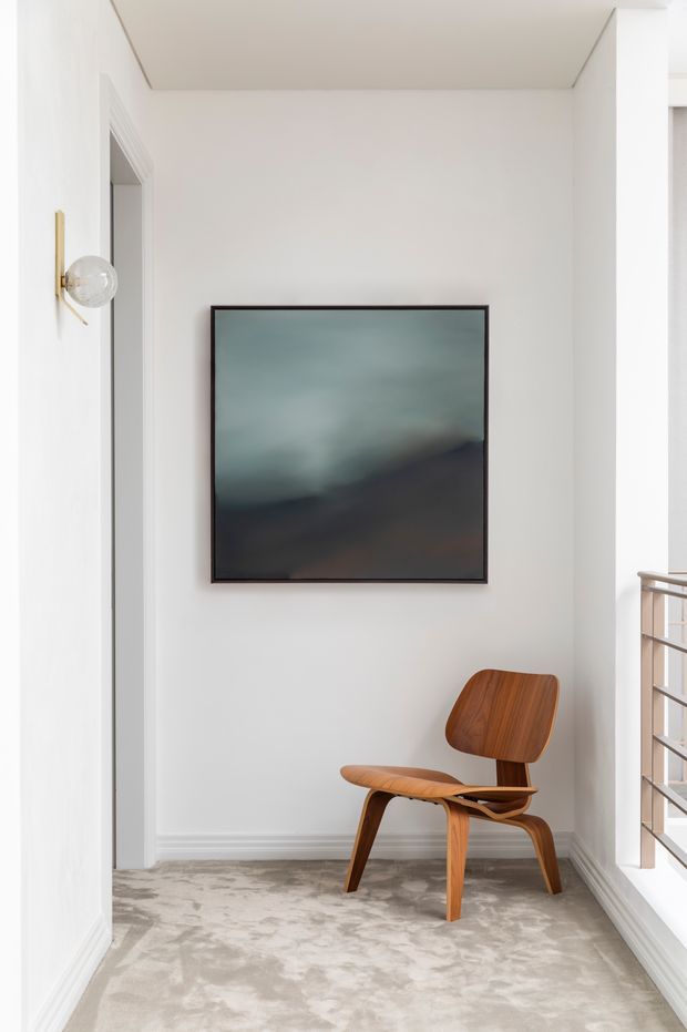 A bright, minimalist landing or hallway nook with white walls and decorative trim, pale patterned carpet, a framed abstract artwork, a sculptural timber lounge chair, and a brass wall sconce. A metal balustrade at right suggests the space is adjacent to a stair or upper-level landing.