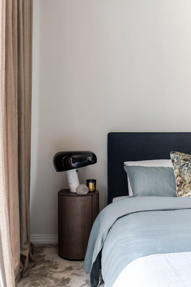 A softly styled bedroom corner featuring a dark upholstered bed with pale blue bedding, a rounded dark wood bedside table, sculptural black-and-white table lamp, and floor-length taupe curtains. The neutral walls and plush carpet create a calm, refined atmosphere.