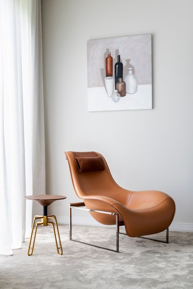 A softly lit minimalist seating corner featuring a sculptural tan leather lounge chair with a matching headrest and polished metal frame, paired with a small round side table with a dark wood top and brass-toned legs. Floor-to-ceiling sheer curtains, pale walls, a light grey carpet, and a neutral still-life artwork create a refined, calm interior.