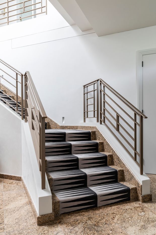 Bright contemporary stairway with terrazzo-clad steps and landings, white walls and ceiling, and slim warm grey metal balustrades with horizontal rails. The staircase is covered by a bold black-and-light patterned stair runner, with a white paneled door visible at right and an upper-level balustrade in the background.
