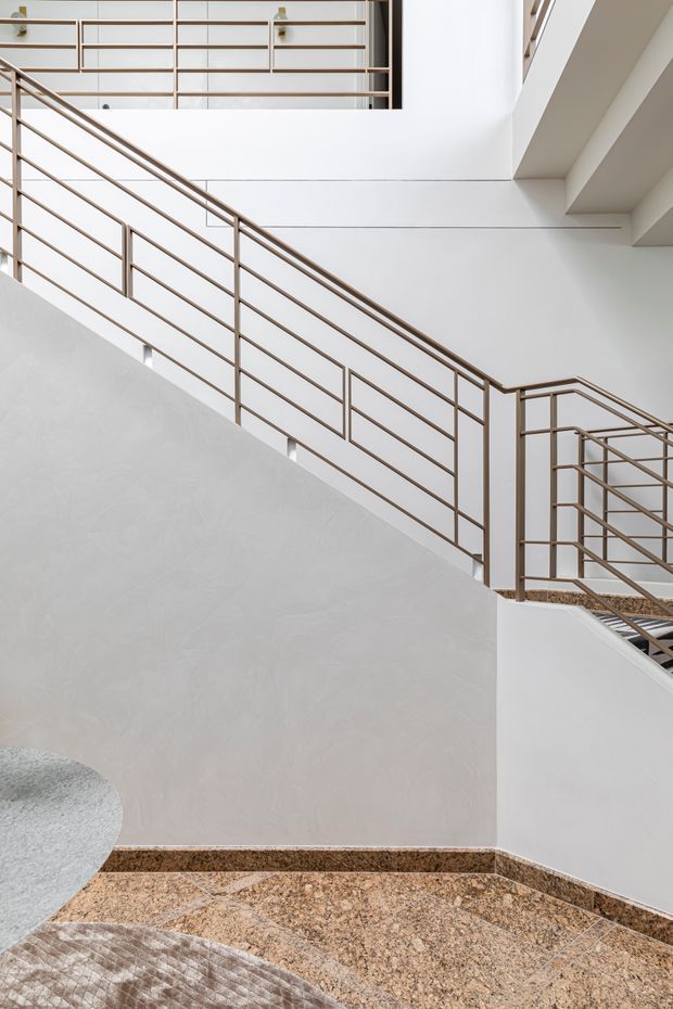 A minimalist multi-level stairway and landing with clean white plaster walls, slender bronze-toned metal balustrades, and a warm speckled terrazzo or stone floor. The composition emphasizes the stair geometry, open void, and layered upper-level walkways, with soft natural light and restrained detailing.