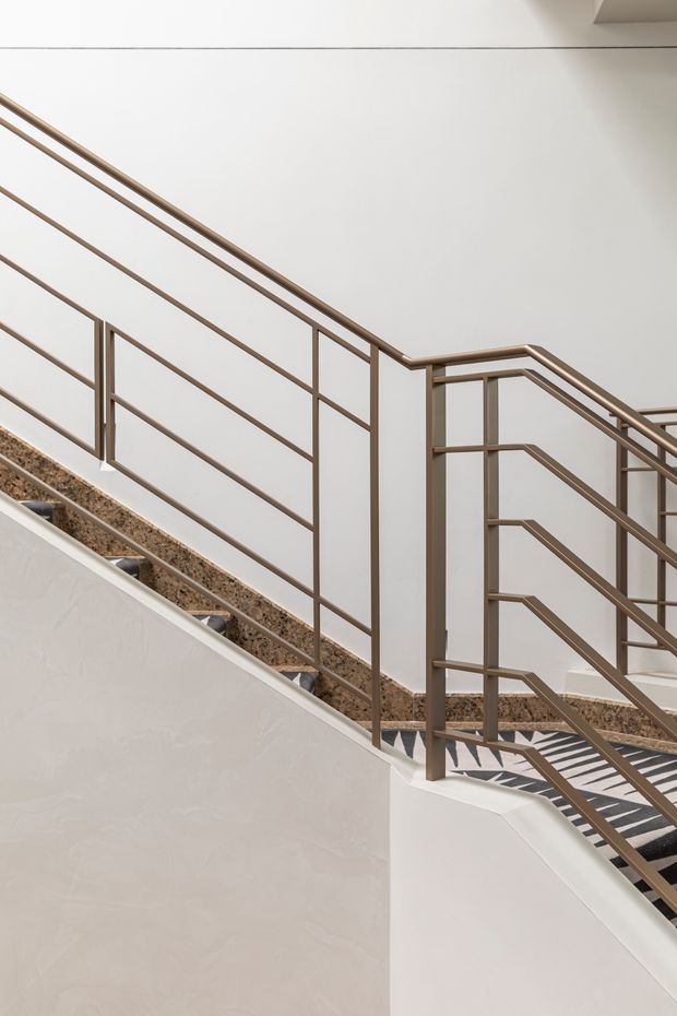 A minimalist interior stairway with warm bronze-toned metal balustrades, white plastered walls, and terrazzo-like stair treads and landings. The stair geometry creates strong diagonal lines, while patterned flooring is visible on the lower landing.