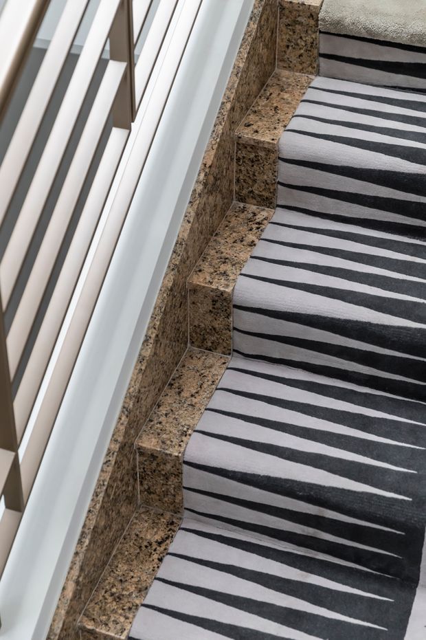 Close-up, angled view of a stairway beside a large window with white framing. The stair treads and risers are finished in speckled brown granite, while a bold black-and-white striped carpet runner covers the steps. The space is brightly lit by the adjacent glazing.