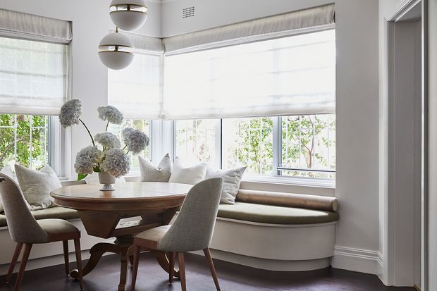 Bright, refined dining nook with a round wooden dining table and upholstered chairs set beside a large bay of windows. A built-in curved window seat with cushions runs beneath the windows, complemented by layered Roman blinds, globe pendant lights, white walls, and dark timber flooring.