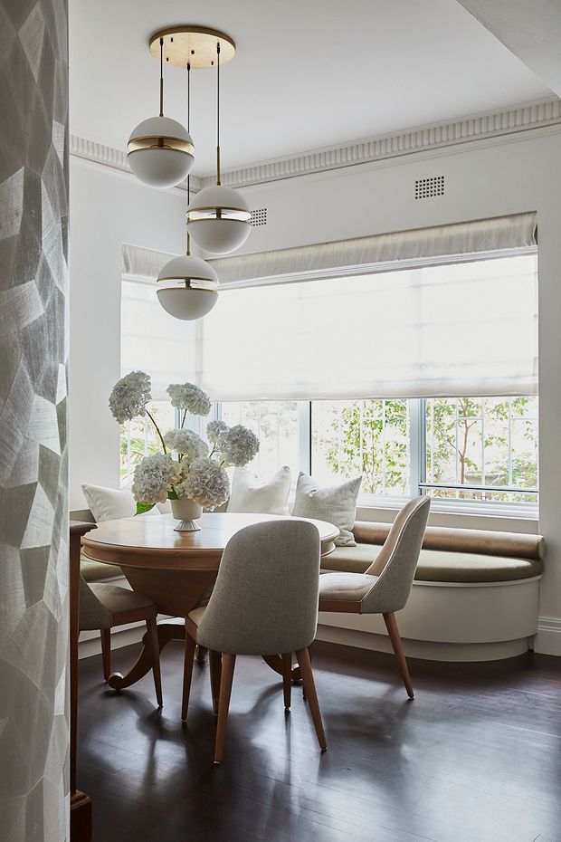 A bright, refined dining nook with a round timber dining table, upholstered dining chairs, a built-in curved window seat, layered cushions, and a large hydrangea arrangement. Floor-to-ceiling-style glazing is fitted with a light roller blind, while sculptural pendant lights hang from a decorative ceiling medallion. The room features dark timber flooring, pale walls with ornate cornice detailing, a geometric textured feature wall, and integrated ventilation grilles.
