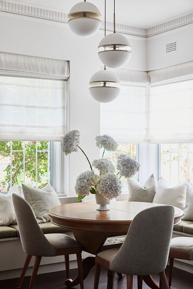 Bright, refined dining nook with a round timber pedestal table surrounded by upholstered dining chairs and a built-in window seat. White upholstered roller blinds, large bay-style windows, decorative ceiling cornices and wall vents create a classic architectural backdrop. Three sculptural white globe pendant lights hang above the table, with a pale hydrangea arrangement as the centrepiece.
