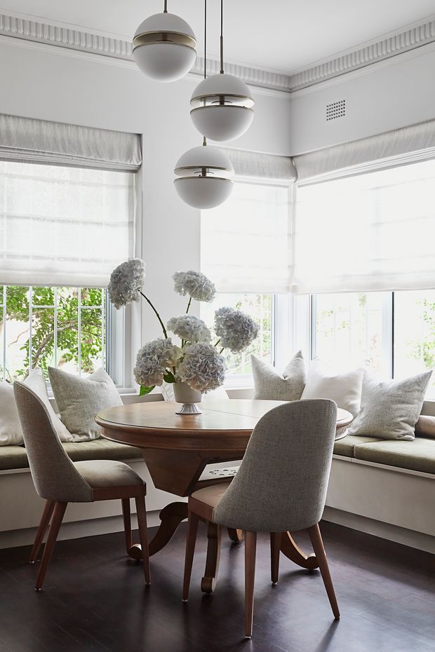 Bright, refined dining nook with a round timber dining table and upholstered mid-century-inspired chairs, surrounded by built-in window seating and large bright windows. The room features white walls, ornate decorative ceiling cornices, layered Roman blinds, dark timber flooring, and a trio of spherical pendant lights. A floral arrangement forms the centrepiece of the table.