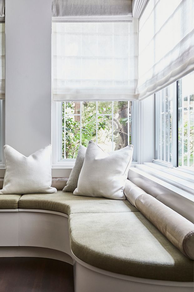 A bright, tailored window-seat nook with a curved built-in upholstered bench wrapping around a corner of large windows. Soft white Roman blinds, pale neutral cushions, light cabinetry, and dark timber flooring create an airy, refined reading or relaxation space.