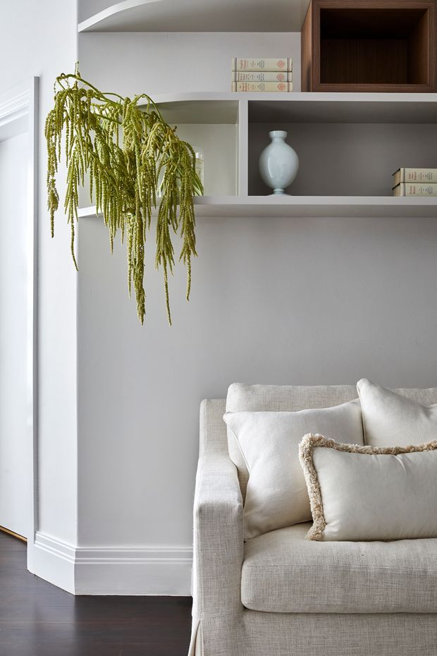 A softly styled living room corner featuring a light beige upholstered sofa with layered cushions, dark timber flooring, pale grey painted walls, white trim, and built-in open shelving. The shelving displays books and a white vase, with a cascading green plant adding an organic accent. The overall look is calm, refined, and contemporary with traditional trim detailing.