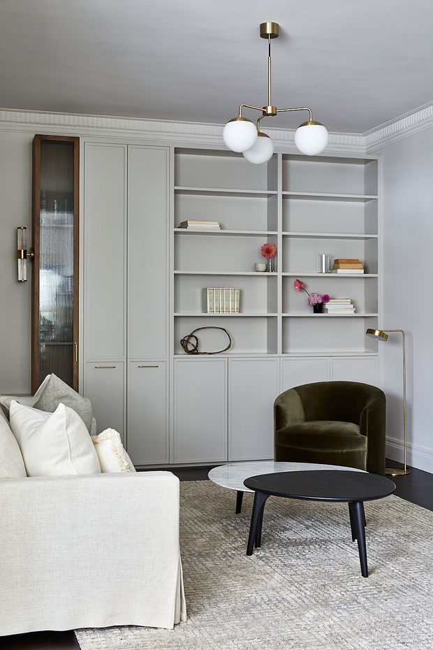Refined living room with custom pale grey built-in cabinetry and open shelving, ornate cornice detailing, a white upholstered sofa, dark olive velvet armchair, layered round coffee tables, and a sculptural brass-and-globe pendant light. The room has a soft neutral palette with dark and brass accents.