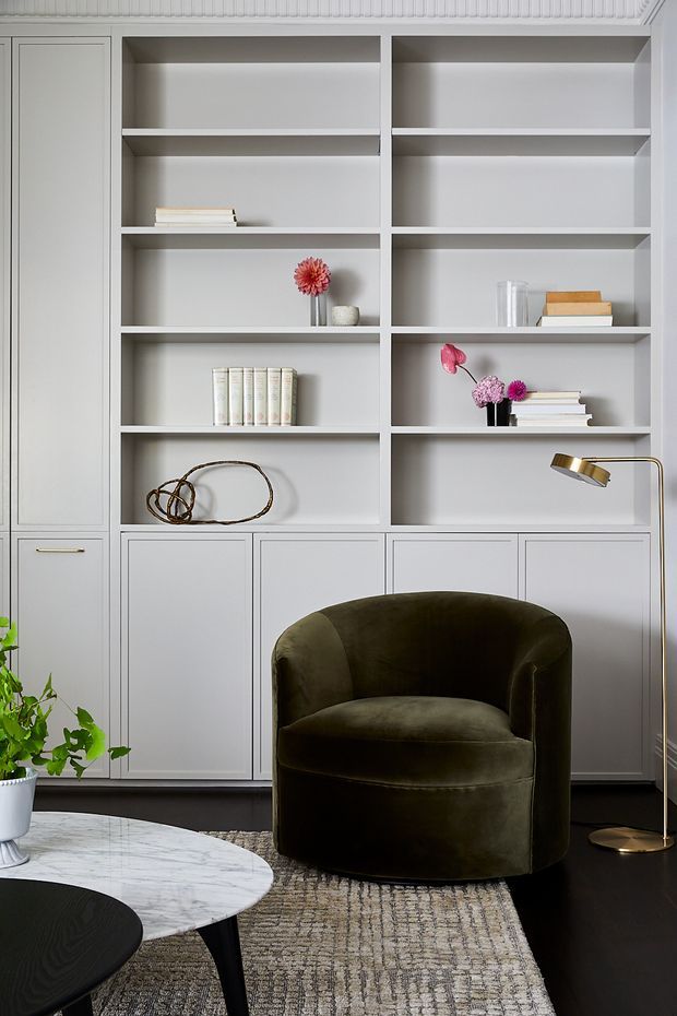 A refined living room corner featuring a full-height built-in white shelving and cabinetry wall with open display niches, books, small floral arrangements and decorative objects. A dark olive velvet barrel chair sits on a textured neutral rug beside a low marble-top and black coffee table, with a slim brass floor lamp. The space has a sophisticated, tailored contemporary-classic feel with strong monochrome contrast and warm metallic accents.