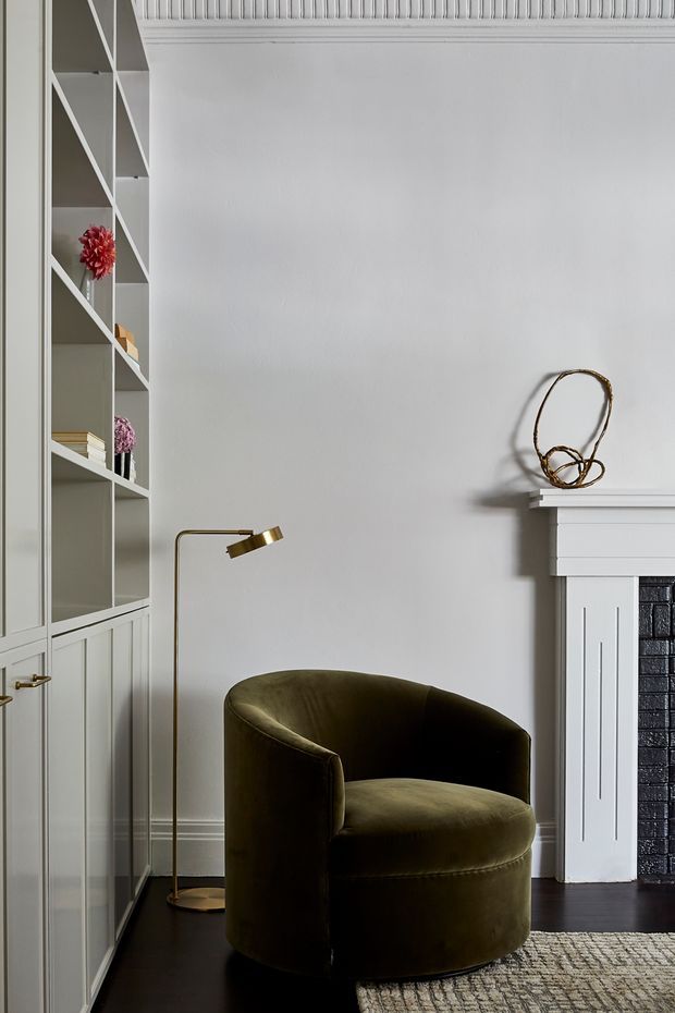 A refined sitting area with a deep olive-green velvet barrel chair positioned beside a slim brass floor lamp. The room features pale grey-white walls, dark timber flooring, built-in floor-to-ceiling shelving and cabinetry with geometric diagonal detailing, a classic fireplace with white mantel and dark brick-lined opening, decorative moulding, and a textured neutral rug. The styling is elegant and restrained with traditional architectural detailing and contemporary furnishings.