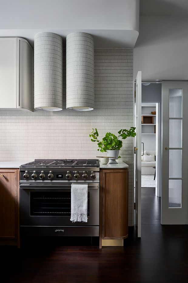 A refined kitchen with a large stainless-steel range cooker, dark timber flooring, warm timber cabinetry, pale vertical mosaic-tile backsplash, and twin cylindrical metal rangehoods. A potted plant sits on the stone benchtop, while glazed internal doors open toward an adjacent room.