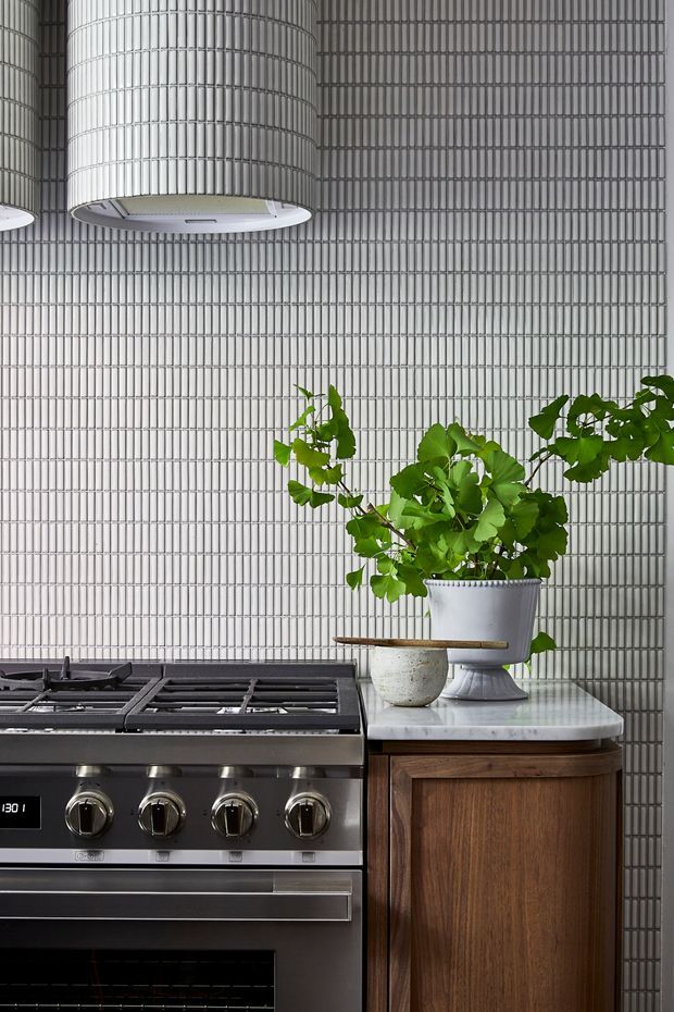 Close-up view of a refined kitchen featuring a large stainless-steel range cooker, a curved timber cabinet with a pale stone countertop, and a dramatic tiled splashback composed of narrow vertical ribbed tiles. A large cylindrical pendant light hangs above, while potted greenery adds a natural accent.