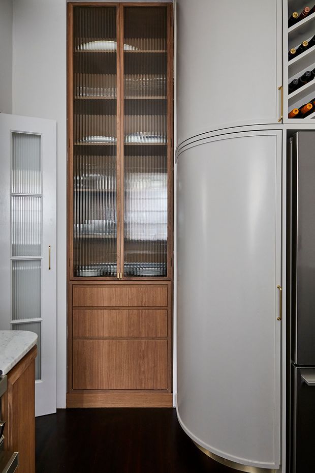 Close-up of a refined kitchen or butler-style storage area featuring a tall walnut-toned built-in cabinet with reeded glass doors, integrated drawers, and adjacent curved painted cabinetry. A wine bottle rack and dark built-in appliances are visible at right, with dark timber flooring and brushed brass hardware.