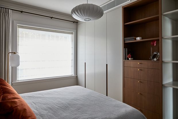 A calm, contemporary bedroom with a low-profile bed in light grey bedding, a warm rust-coloured cushion, a large window with a simple roller blind, and integrated floor-to-ceiling wardrobes. A dark timber built-in shelving and drawer unit provides storage and display space, while sculptural pendant lighting and a slim floor lamp add understated detail.