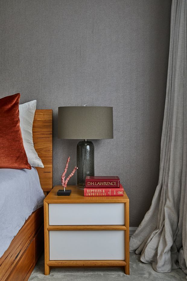Close-up of a stylish bedroom corner featuring a timber bed frame and bedside table with pale inset drawer fronts, a dark textured table lamp, books, and a small decorative vase. The room has grey textured walls, grey carpet, floor-length grey curtains, and layered bedding with a rust-red accent cushion.