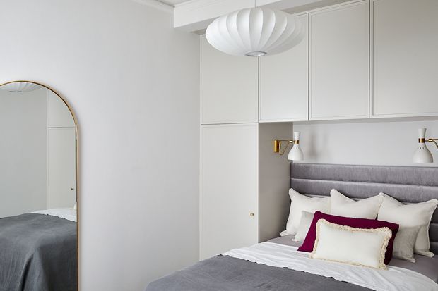 A refined bedroom with a grey upholstered bed and layered neutral bedding, including a burgundy accent cushion and cream decorative pillow. Full-height built-in cabinetry surrounds the headboard, with integrated bedside niches and small wall lights. The room features light grey wall panelling, white decorative cornices, a large arched mirror with a slim brass frame, and a sculptural white pendant light.