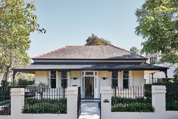Front exterior view of a single-storey period-style residence with a broad hipped tiled roof, corrugated metal verandah awning, cream rendered walls, dark window and door trim, and a formal front garden enclosed by a low wall and iron fencing.