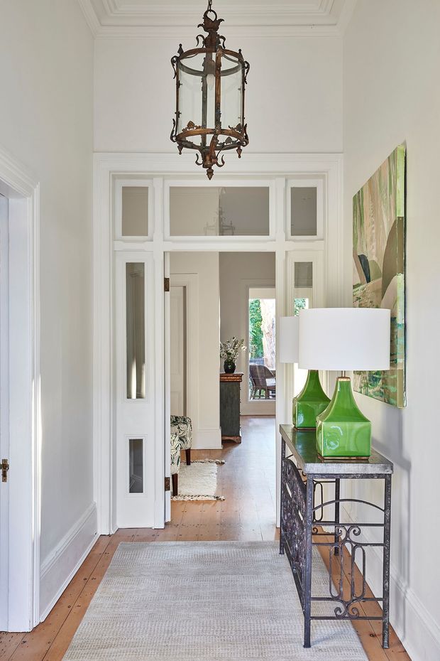 Bright, narrow traditional hallway with white painted walls, ornate moulded trim, panelled door surrounds, timber floorboards and a pale runner. A decorative antique-style lantern pendant hangs from the coffered ceiling. On the right is a dark wrought-metal console table with green glass lamps, while an abstract artwork provides a colourful accent. The hallway opens toward a further sitting or living area with a glimpse of outdoor greenery.