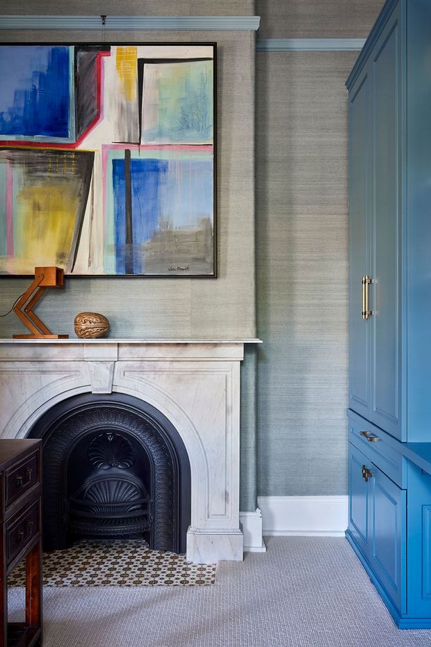 A refined traditional interior vignette featuring a white marble fireplace with an ornate black cast-iron insert, patterned hearth tiles, textured grey wallcovering, pale carpet, and a vivid blue built-in cabinet with brass hardware. A colourful abstract artwork and small decorative objects sit above the mantel, creating a classic yet contemporary layered look.