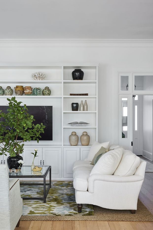 Bright, refined living room with white built-in cabinetry and open display shelving surrounding a television, layered with decorative ceramics and greenery. A plush white sofa, glass-and-metal coffee table, patterned area rug, and pale timber flooring create a calm, classic interior. A glazed doorway and adjacent circulation space are visible to the right.